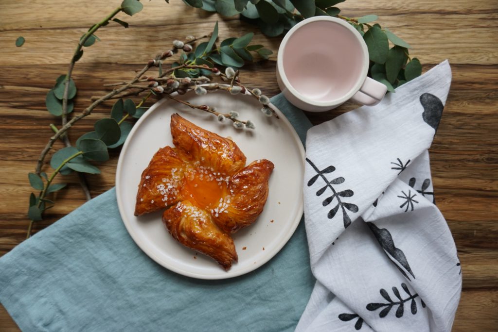 A baked good sits on a ceramic plate next to some greenery, a mug, and handmade block printed linen napkins