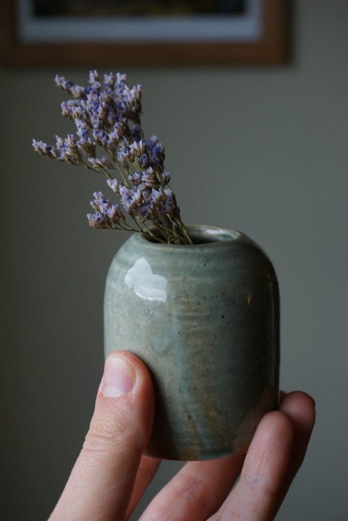 A hand holds up a small vase with purple flowers in it