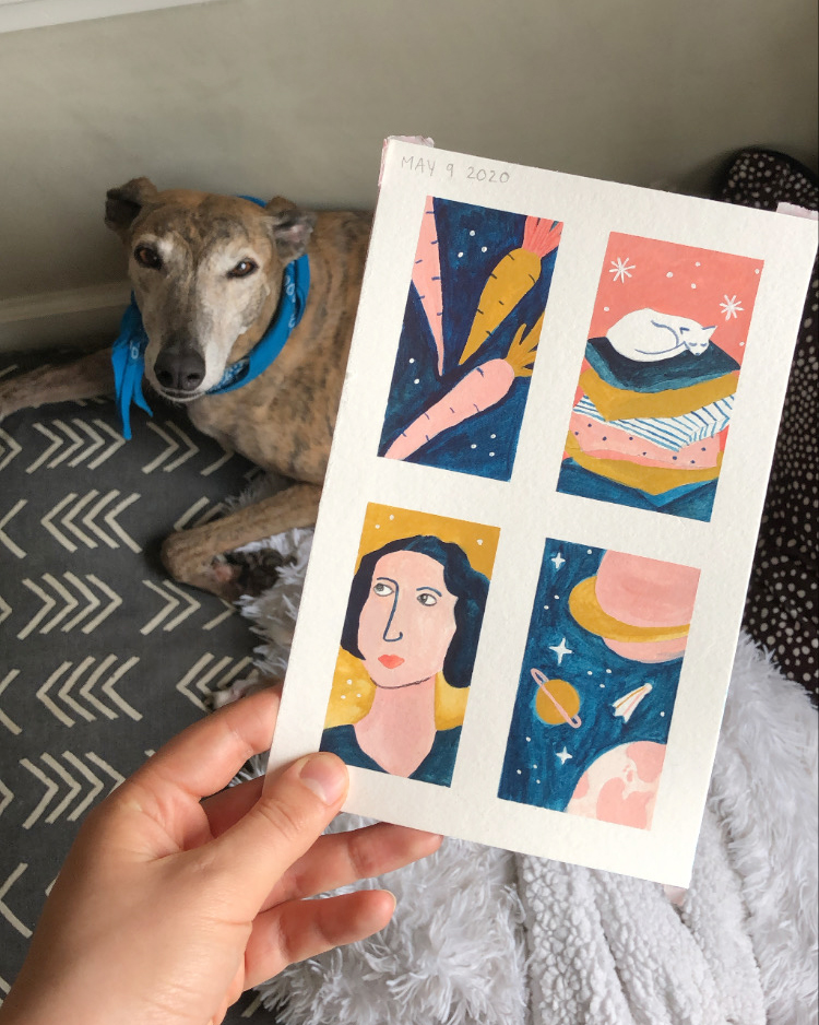 Four small paintings of carrots, a napping cat, a woman's face, and a space scene being held up in front of Greer the greyhound